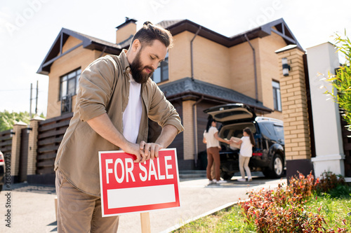 Foto Young bearded father placing For sale sign into ground near house while putting