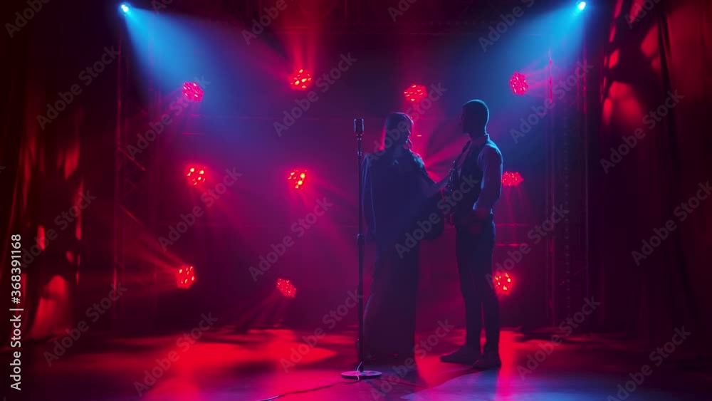 Silhouette a young girl singer looks passionately at a guy saxophonist and touches it with his hand then sings a song on stage in a vintage microphone. A brunette in a long black dress and men jacket
