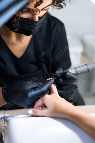 manicurist master in black gloves and mask is making manicure with a manicure drill apparatus