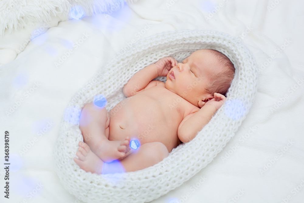 A cute adorable newborn baby wrapped in a white knitted cocoon sleeps with its legs bent in the Lotus position.  Blurred blue garlands ahead.