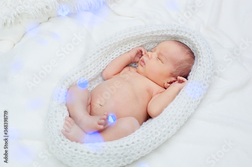 A cute adorable newborn baby wrapped in a white knitted cocoon sleeps with its legs bent in the Lotus position. Blurred blue garlands ahead.