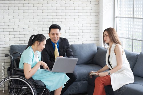 Young disabled woman sit on wheelchair show her presentation on a laptop and discuss with her two colleagues in the workplace office room.