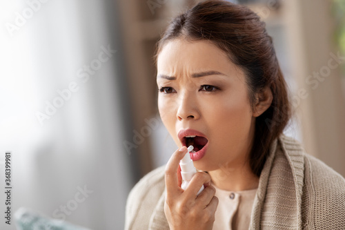 health, cold and people concept - sad sick young asian woman in blanket using oral spray medicine at home
