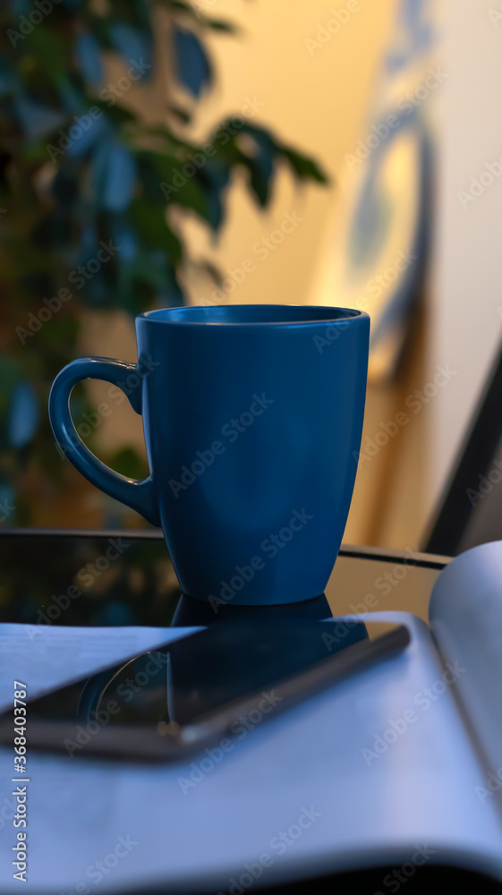 Evening coffee. Blue cup with coffee, open book and mobile phone, evening light, sunset, dusk. Atmospheric, cozy evening in the garden, green leaves, conversation. Warm light, blur bokeh. Evening coffee. Blue cup with coffee, open book and mobile phone, evening light, sunset, dusk. Atmospheric, cozy evening in the garden, green leaves, conversation. Warm light, blur bokeh.