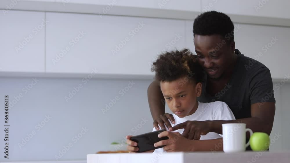 Young father and son are using smartphone while sitting at table in home kitchen spbi. African American man and boy looking at screen with smiles and holding device in hands at desk in light interior Young father and son are using smartphone while sitting at table in home kitchen spbi. African American man and boy looking at screen with smiles and holding device in hands at desk in light interior