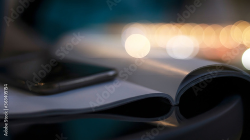 Open book and phone on a glass black table. Blurred boke background, close-up. Dark evening background with sunset highlights.