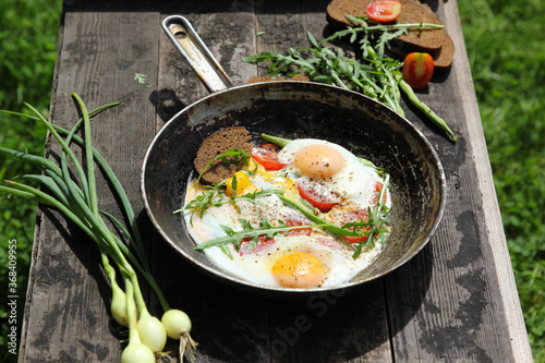 Rustic breakfast. Fried eggs with vegetables: tomatoes and asparagus beans in a pan with bread, herbs, onions on old wooden black background in nature. Summer, sun light. Background image, copy space