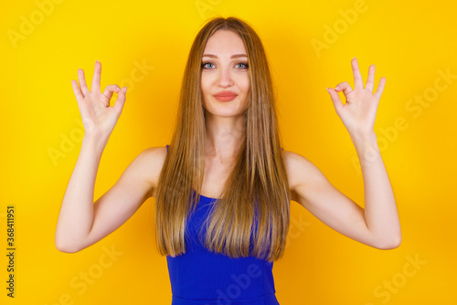 Glad attractive woman shows ok sign with both hands as expresses approval, has cheerful expression. Photo of beautiful female has appealing appearance, being optimistic. Standing against gray wall.