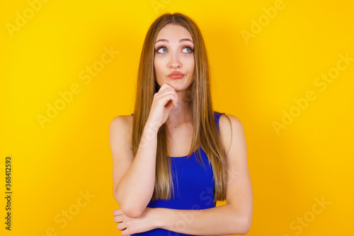 Portrait of thoughtful woman keeps hand under chin, looks away trying to remember something or listens something with interest, dressed casually, poses indoors. Youth concept.