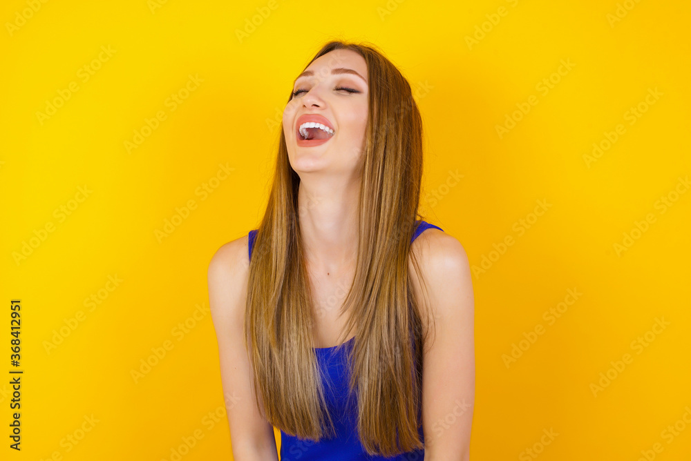 Joyful European woman having fun and laughs at good joke or sings wears casual clothes, standing against gray wall. Happy woman with long hair poses inddoors. Joyful European woman having fun and laughs at good joke or sings wears casual clothes, standing against gray wall. Happy woman with long hair poses inddoors.