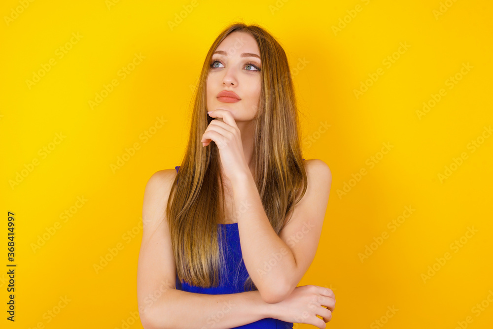 Portrait of thoughtful smiling girl keeps hand under chin, looks sideways, thinking or wondering about something with interest, dressed casually, poses against gray studio. Taking decisions concept. Portrait of thoughtful smiling girl keeps hand under chin, looks sideways, thinking or wondering about something with interest, dressed casually, poses against gray studio. Taking decisions concept.