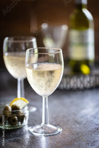 2 glasses of chilled sparkling white wine with dish of olives and sliced lemon. Drinks tray with wine bottle and glass in background