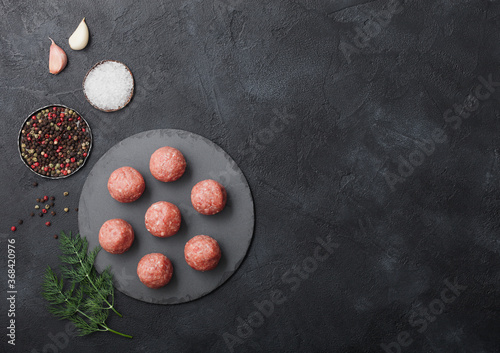 Fresh raw minced beef meatballs on round board with pepper, salt and garlic on black background.