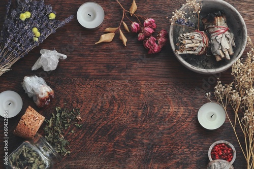 Dark brown wooden table background for text, decorated with witchy wiccan items to look esoteric and occult. Free writing space in the middle. Dried herbs, crystals, sage smudge sticks candles flowers photo