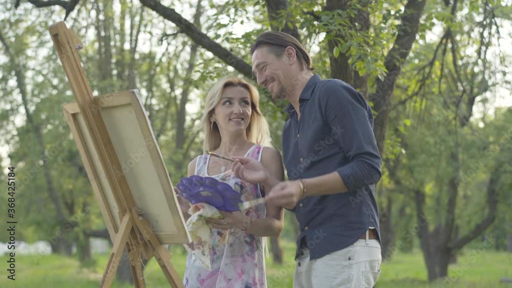 Side view of positive Caucasian man and woman discussing process of picture painting outdoors. Smiling Caucasian couple creating artwork together in park. Creativity and love concept. Side view of positive Caucasian man and woman discussing process of picture painting outdoors. Smiling Caucasian couple creating artwork together in park. Creativity and love concept.