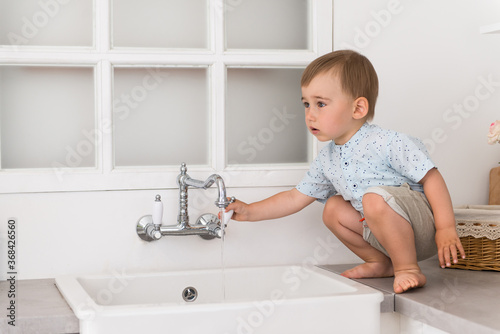 portrait of a child in home kitchen - baby sits near washbasin and opens water