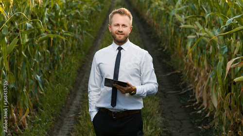 Mature, handsome, well-dressed, businessman holding a new tablet, smiling and standing in the middle of green and yellow cornfield.