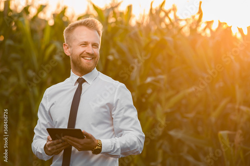Adult, handsome, stylish, blonde, businessman holding a new tablet and standing in the middle of green and yellow corn field during sunrise.