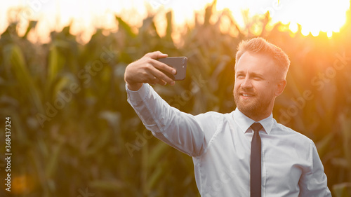 Stylish, blonde, businessman taking a photo in the middle of green and yellow cornfield during sunrise.