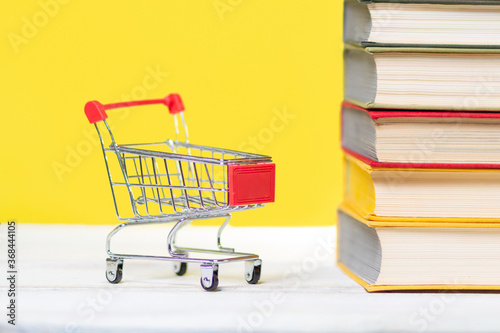 Back to school. A mini shopping cart and a stack of books with an open book at the top on a yellow background. Concept of education, reading and buying books