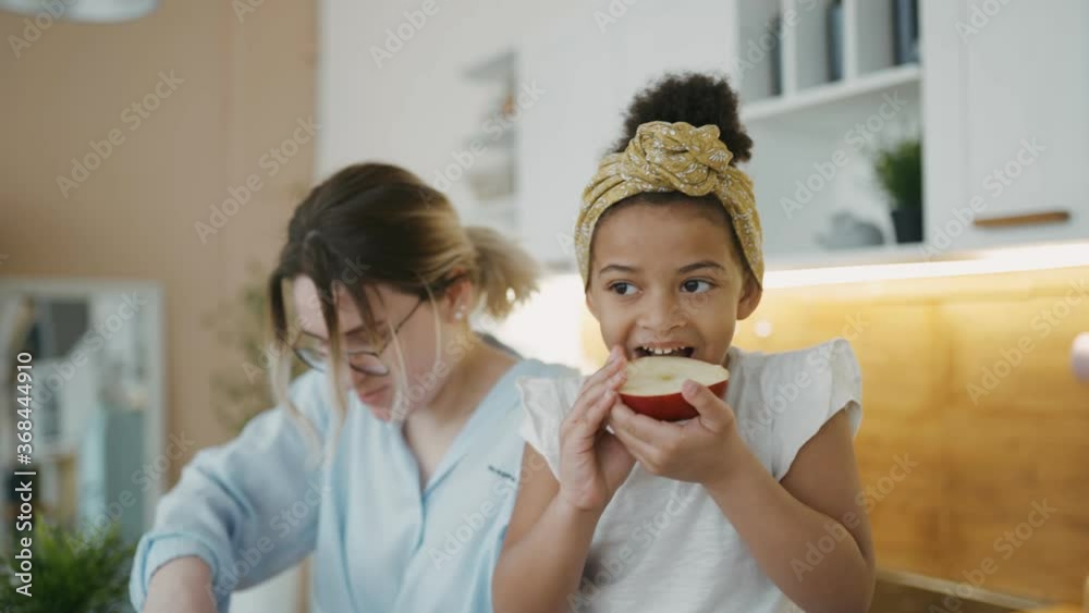 Snack in kitchen at home. Portrait of beautiful cute little African girl sitting on table biting red apple. In background, Mom's making lunch. Multiracial family, healthy home-cooked food, cooking. Snack in kitchen at home. Portrait of beautiful cute little African girl sitting on table biting red apple. In background, Mom's making lunch. Multiracial family, healthy home-cooked food, cooking.