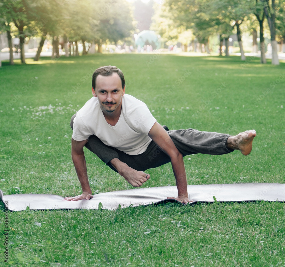 Fototapeta premium attractive smiling young caucasian man doing hatha yoga exercises in city park in summer sunny time. Outdoor Asanas
