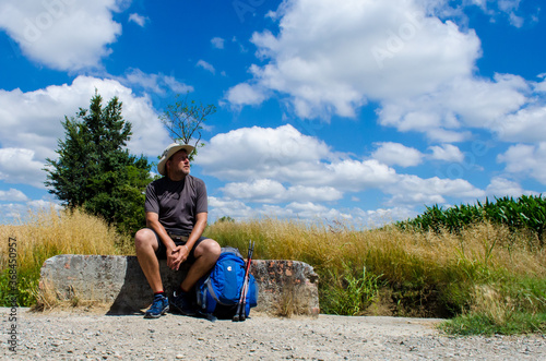 Un pellegrino si riposa lungo la Via Francigena con alle spalle la campagna e un bel cielo azzurro con delle nuvole photo