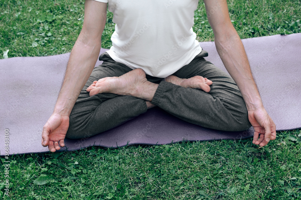 Fototapeta premium young man doing yoga on green grass, lawn in city park in summer sunny time. A man sits on a mat, meditates, hands folded in mudras