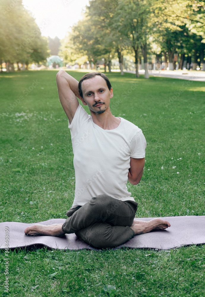 Fototapeta premium young man doing hatha yoga on green grass, lawn in city park in summer sunny time. A man sits on a rug, meditates, hands folded behind his back