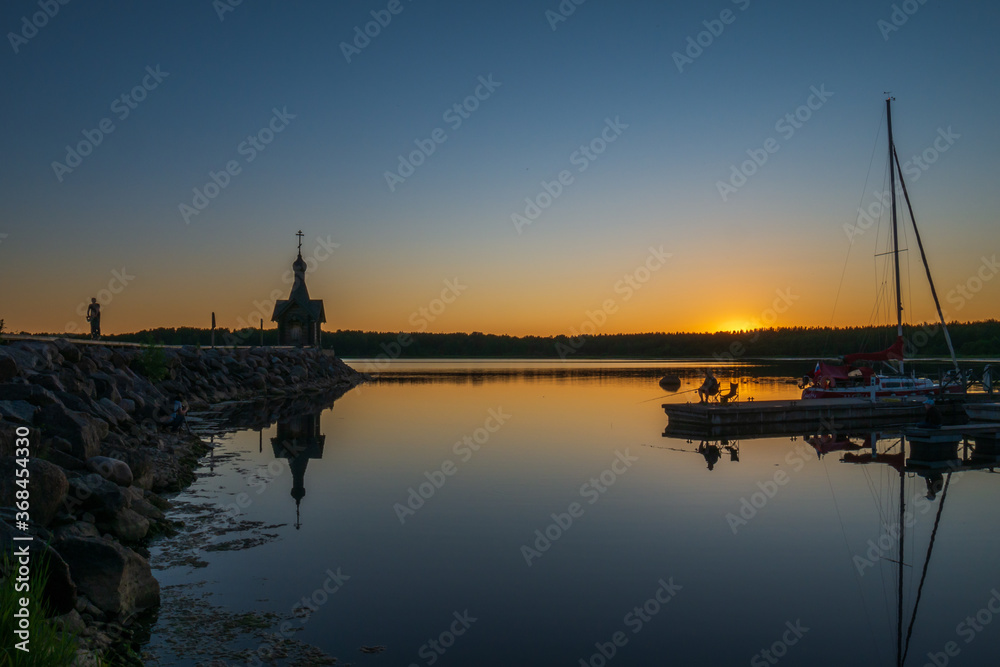 The silhouette of a church, a fisherman and a boat against the backdrop of the sunset. Night, reflection in the water. Dark silhouettes of the chapel.
