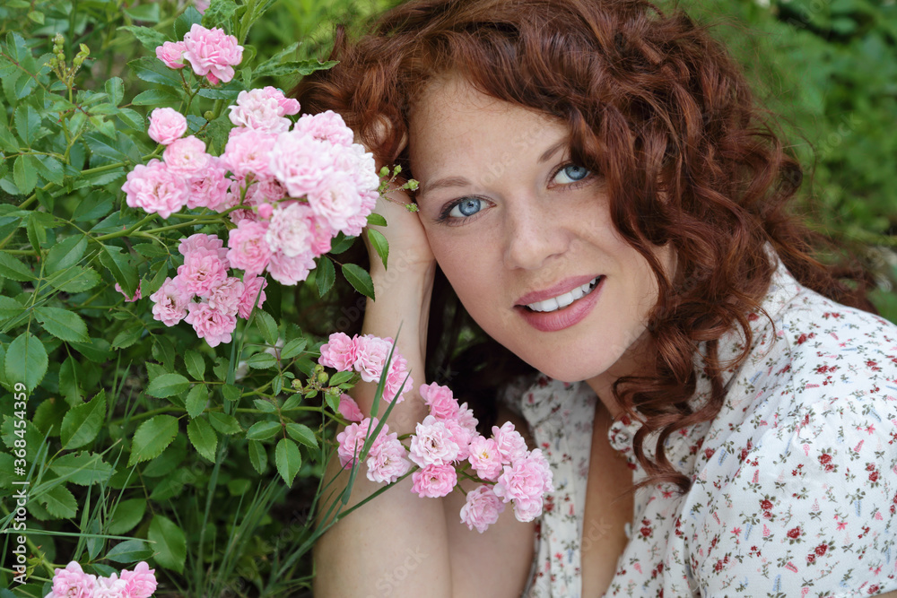 portrait naturel d'une jeune et jolie femme rousse aux long cheveux bouclés souriante dans la nature avec des fleurs roses en été 