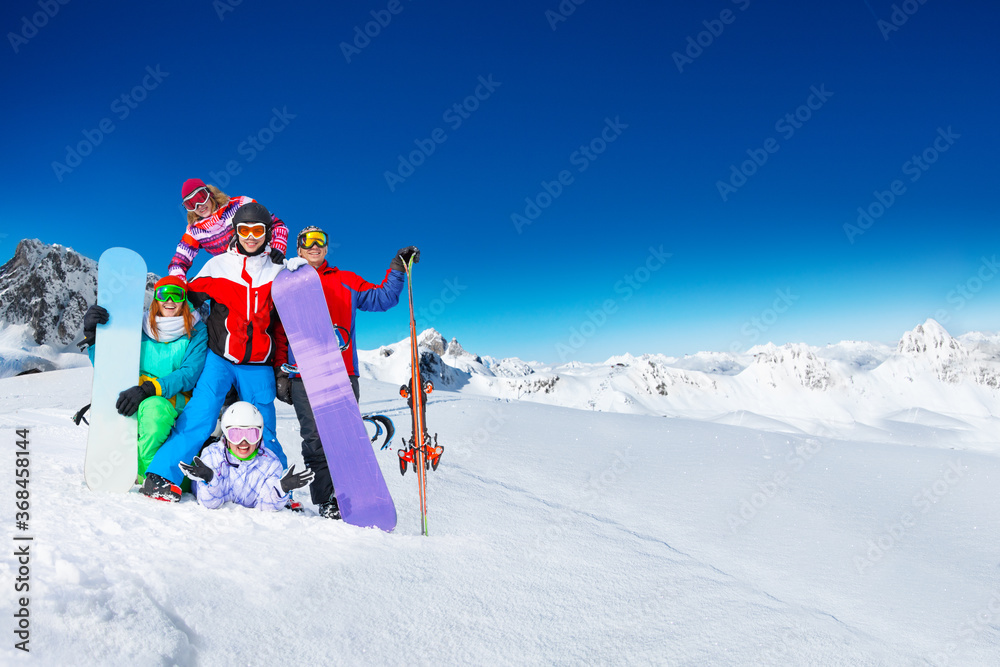 Group of young adults snowboarders pose together on top of the mountain over snow covered peaks on sunny weather on back