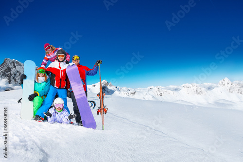 Group of young adults snowboarders pose together on top of the mountain over snow covered peaks on sunny weather on back