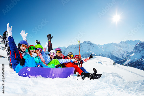 Group of happy young adults hold snowboard sit together with lifted hands waving