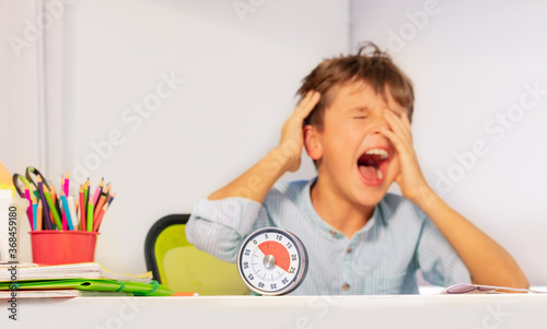 Sad crying autistic boy sitting during development therapy by the desk with lesson timer cover face using hands