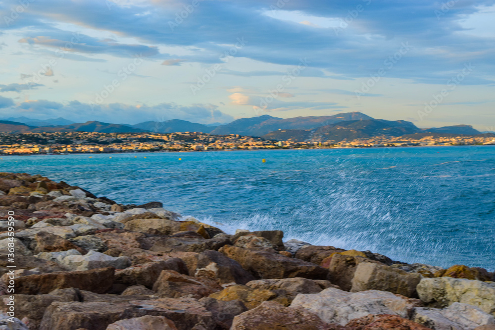 Waves crashing on the rocky breakwater against the backdrop of the Mediterranean sea and the Nice city background. France.