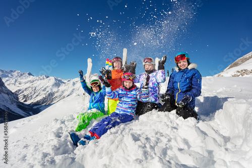 Fun group portrait of kids sit together throw snow in the air over beautiful mountain range tops in Alps
