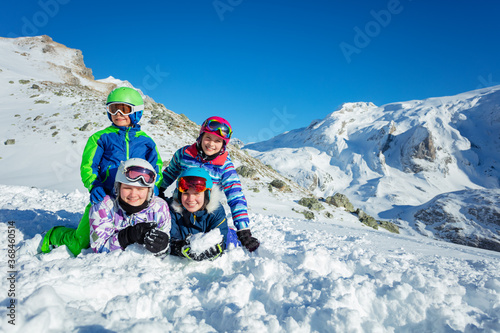 Group of four smiling happy kids lay together in the snow on mountain top wear ski outfit, color helmets and glasses