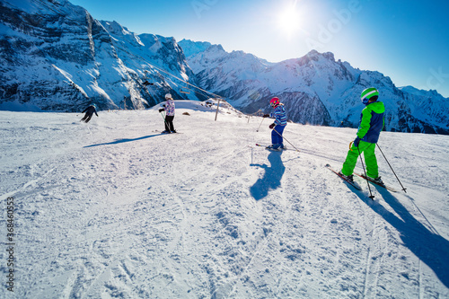 Four kids ski downhill in Alpine slope in school formation together one after another view from behind with mountains on background