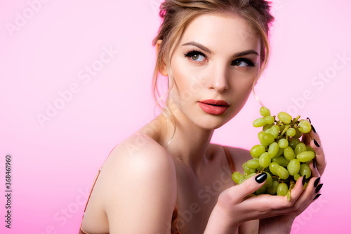 elegant beautiful blonde woman holding green grapes and looking away isolated on pink