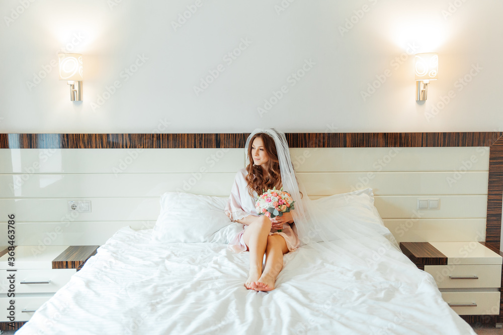 Morning of the bride in a pink robe and a veil in the hotel on the bed, wedding dress Morning of the bride in a pink robe and a veil in the hotel on the bed, wedding dress