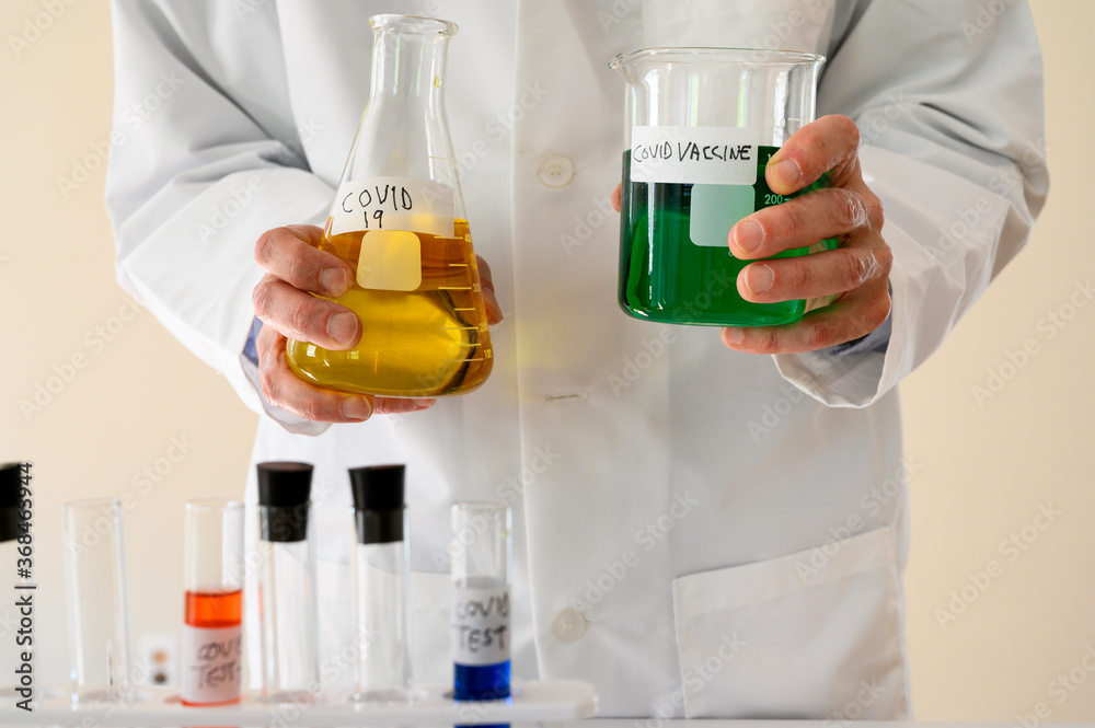 Covid-19 Reseracher in a lab coat holds a filled flask and beaker. Test tubes in the foreground. Covid-19 Reseracher in a lab coat holds a filled flask and beaker. Test tubes in the foreground.