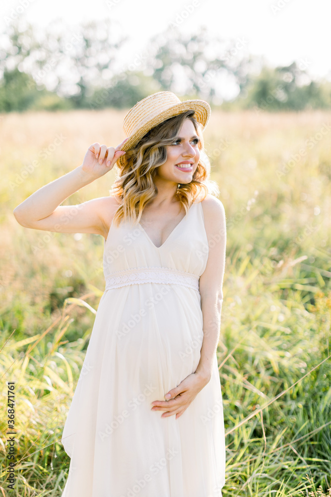 Smiling charming young pregnant woman, wearing white dress and straw hat, resting in summer field. Posing outdoors. Motherhood. Outdoor portrait of beautiful pregnant woman in white dress Smiling charming young pregnant woman, wearing white dress and straw hat, resting in summer field. Posing outdoors. Motherhood. Outdoor portrait of beautiful pregnant woman in white dress