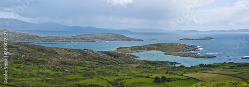 Derrynane Bay, Kerry, Ireland. Rocky islands in a blue sea with distant mountains visible through rain showers, and a rugged pasture of green fields and stone walls in the foreground.