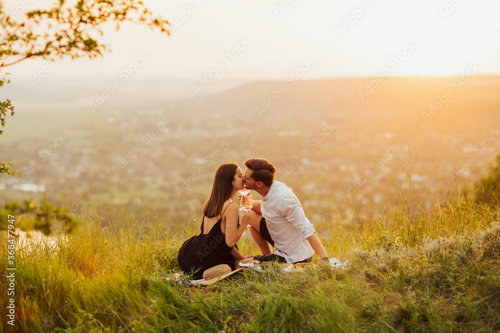Happy gorgeous elegant couple kissing on summer picnic. They are relaxing together on a summer day. Couple in love on a white plaid on mountain at a picnic against the background of the sunset