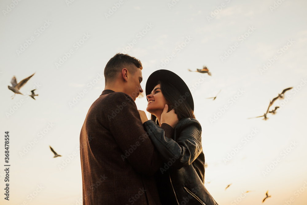Happy young romantic couple hugging and looks at each other with love. Young couple sharing happy and love mood on the beach with flying seagulls on background.