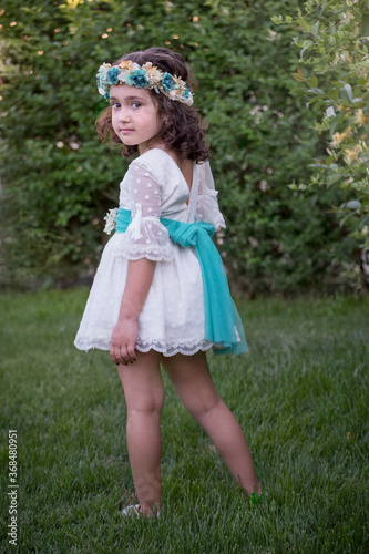 Girl in a white dress with green bow posing in a garden. Curly brown hair.