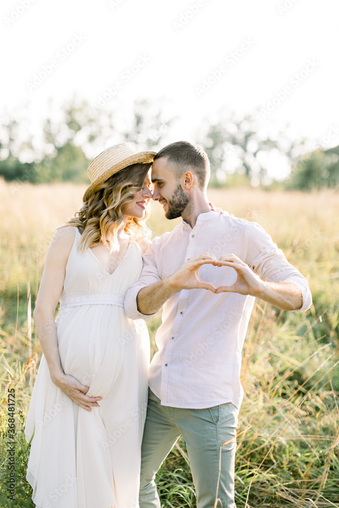 Young husband and his pregnant wife in hat and white dress, walking in the sunny summer field. Man makes a heart gesture with his hands and looks at his woman