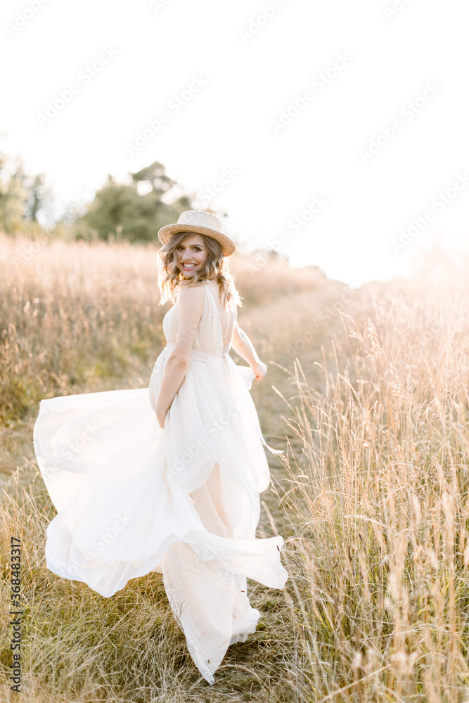 Smiling charming young pregnant woman, wearing white dress and straw hat, resting in summer field. Posing outdoors. Motherhood. Outdoor portrait of beautiful pregnant woman in white dress