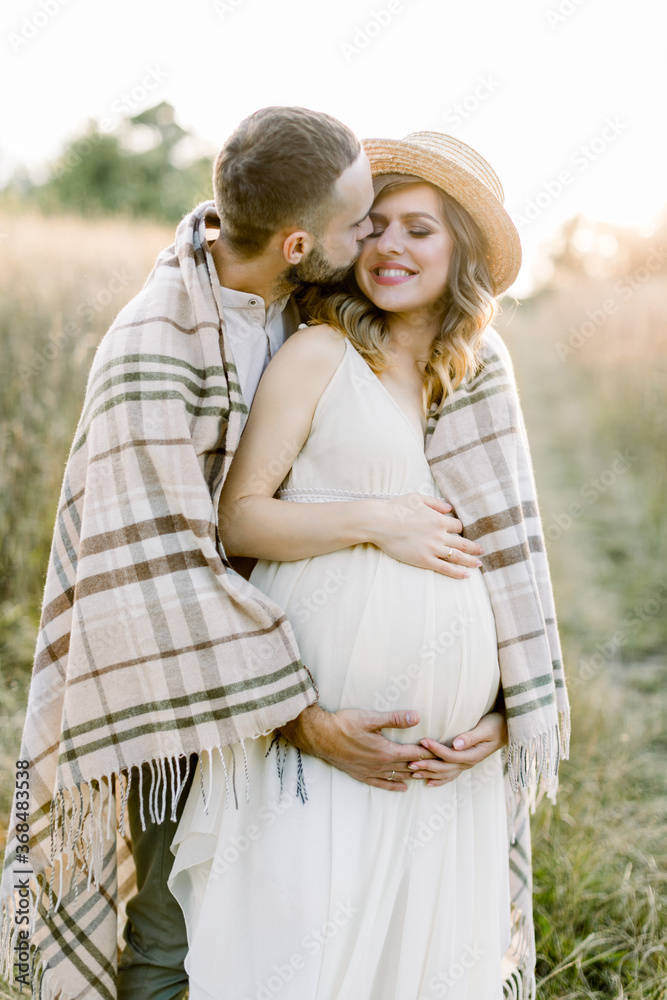 Fototapeta premium Handsome Caucasian man hugging his attractive pregnant woman in straw hat and white dress in sunny day in field. Happy couple covered with blanket, waiting for a baby